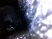 Close-up of an intake valve inside an engine. The valve face is dark and rough with heavy black carbon buildup and oily residue, while the metal edges appear bright and clean. The surrounding area is blurred and dark, suggesting a technical engine inspection view. The scene feels serious and maintenance-focused, showing a valve that needs cleaning. No readable text is visible.
